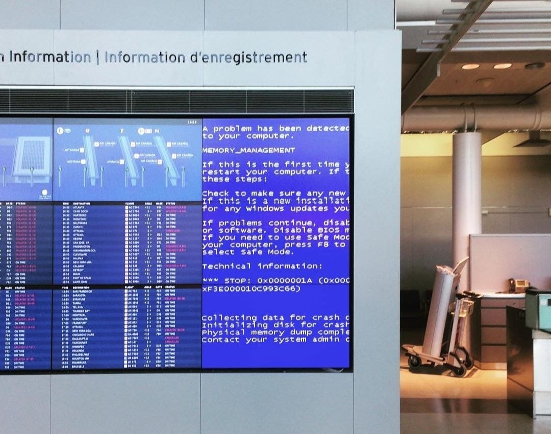 A Canadian airport departures board showing a Windows blue screen of death (MEMORY_MANAGEMENT error) on one of its display panels, next to the flight status listings
