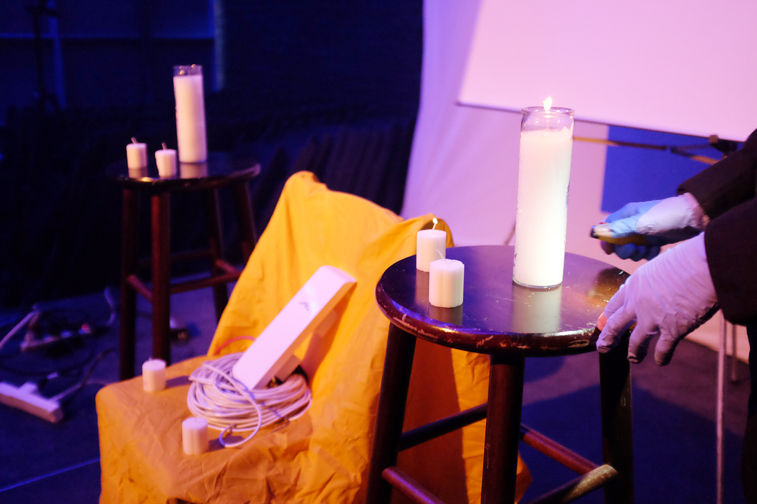 A router and ethernet cables sit on a chair draped with yellow cloth. Beside it are two small tables with white pillar candles. Behind is a whiteboard. Towards the right of the image, there are two hands wearing latex gloves